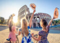 Three happy young women friends tourists with bikes waving hats at Colosseum in Rome, Italy at sunrise.