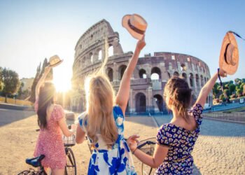 Three happy young women friends tourists with bikes waving hats at Colosseum in Rome, Italy at sunrise.
