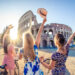 Three happy young women friends tourists with bikes waving hats at Colosseum in Rome, Italy at sunrise.
